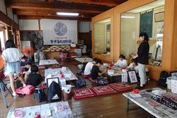 大きなショーケースが設置されたフローリング床の室内で、引率の先生が立って見ているなか、様々な道具や材料が置かれているテーブルの上で子供たちが座布団に座って絵付け体験をしている様子の写真