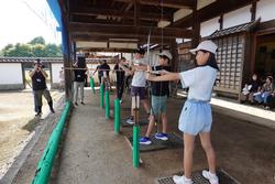 大きな屋根のある屋外の弓道場で、一列に並んだ数人の子供たちが的に向かって真剣な表情で弓を構え弓道体験をしている写真