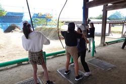 大きな屋根のある屋外の弓道場で、他の女の子が弓を持って見ているなか、先生らしき女性が女の子に弓の構え方を教えている写真