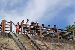 小高い場所にある階段を上がった天文台の上で、リュックを背負った複数の子供たちが笑顔でカメラに向かって手を振ったり、ポーズをとったりしている写真