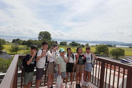 後ろに、木々に囲まれた広大な田んぼ、湖、そして遠くの山々が見渡せる展望台のような場所で、数人の子供たちが横一列に並んでカメラに向かってポーズを取っている写真