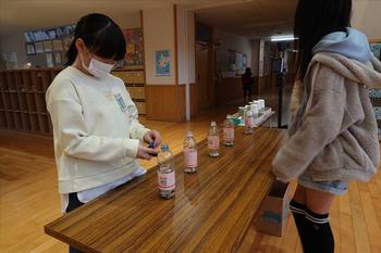 机に並べられたペットボトルの募金箱に女の子2名が向かい合って立っている写真