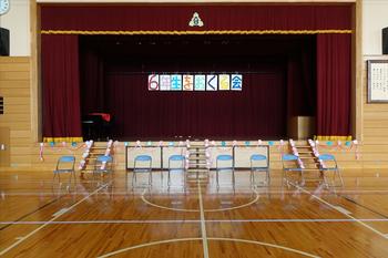 背景の赤い幕に「6年生を送る会」と書かれたカラフルな文字が掲げられ、ピアノが設置された体育館内のステージの縁や左右の階段が青とピンク色の手造りの花で装飾され、ステージ下に8個のパイプ椅子が並んでいる写真