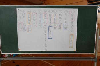 大きな模造紙に書かれた「6年生を送る会」の進行表が黒板に貼られており、各学年の発表内容や順番が確認できる写真