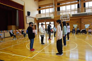体育館の前方で児童たちが向かい合って立ち、左側に立っている児童がプレゼントの様な物を両手で持っている場面の写真