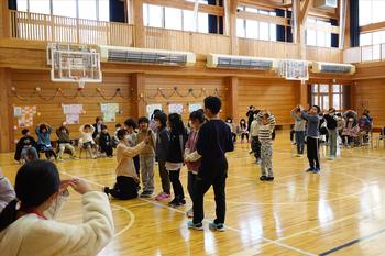 体育館の中央で他の子供たちが踊っているなか、6人の児童が横一列に並んで立ち、左から3番目の児童の前に膝まづいた女の子がマイクを向けている様子の写真