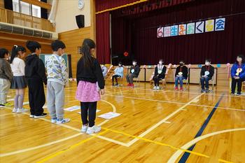 ステージ前に設置されたイスに座る6年生に向かって発表している横一列に並んだ低学年の児童たちの写真