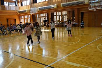 体育館の中央で4人の女の子たちが横一列に並んで手を握り、ステップを踏んでダンスをしている様子の写真