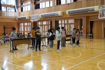 体育館の後方で椅子に座った他の児童たちが見ているなか、数名の児童が木琴や鉄琴、鍵盤などの楽器で演奏を披露している様子の写真