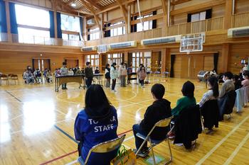 楽器を演奏している下級生たちを、6年生たちが椅子に座って見ている様子を後方から撮影した写真