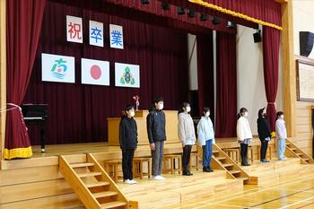 背景に一文字ずつ「祝 卒業」と書かれた幕と国旗や町旗、校旗が掲げられた体育館のステージ前の段の上に、横一列に並んだ7人の児童たちが卒業式の練習を行っている様子の写真