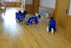 青いジャージを着た複数の児童たちが、廊下の床に膝をついて拭き掃除を行っている様子の写真