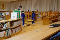 周りに本棚や椅子をあげた勉強机が並んでいる図書館内で、青いジャージを着た複数の子供たちがモップをかけて掃除をしている様子の写真