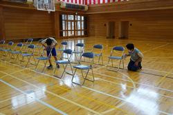 バスケットゴールのある体育館中央に並んだ2列のパイプ椅子を、2人の子供たちが拭いている写真