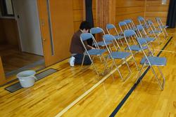 バケツが置かれた体育館入り口後方の床に、たくさんのパイプ椅子が並べられ、一人の児童がしゃがんで椅子の脚を拭いている様子の写真