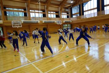 学校の体育館で、お揃いの青いジャージ姿の生徒たちが、両手と両足を横に広げてよさこいの練習をしている写真