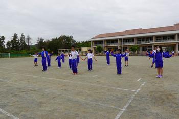 奥に校舎が見える校庭で、両手を広げて間隔をあけて立っている青と白のジャージを着た生徒たちが、よさこいの練習を行っている写真