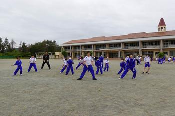 奥に赤い屋根の校舎が見えるグラウンドで、間隔をあけて両足を広げて立つ、お揃いの青と白のジャージ複数の生徒たちが、黒いジャージ姿の先生と一緒によさこいの練習をしている写真