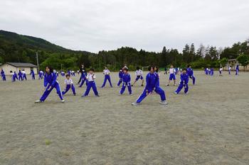 山や木々に囲まれた学校の校庭で、両足を横に大きく広げて間隔をあけて立っている青いジャージを着た大勢の生徒たちが、よさこいの踊りの練習をしている写真