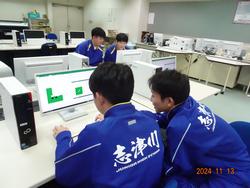 白いデスクトップ型コンピュータを複数置いたテーブルが並んだ室内で、二人一組でモニターを見ながら作業をしている男子生徒たちの写真