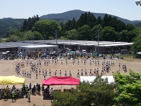 山に囲まれた小学校の校庭で赤色や黄色のテントが並び校庭に赤白帽を被り体操服姿の児童が2重に円形を描いて演技をしている様子や、奥には仮設住宅が並んでいる写真