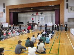 体育館の前方に先生が横一列に並び床に体育座りをして話を聞いている児童の写真