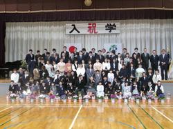 入学と書かれた横断幕が飾られた舞台の前に椅子が並べられ一年生が座り後ろに保護者が立った記念撮影の写真