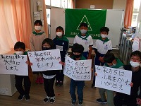 小学校の教室で、4人の子供たちがそれぞれ手にスローガンが書かれた紙を持ってカメラ目線で立っている様子の写真