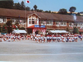 国旗が掲揚され白いテントが設置された校庭で、大勢の子どもたちが校舎前に集まり賑やかな運動会の様子の写真