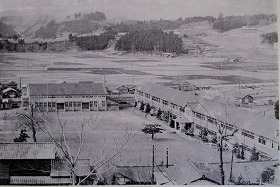昔の木造校舎と広い運動場、背後に田畑と山並みが広がるモノクロ写真