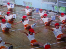 等間隔に並んだ赤白帽子に体操服姿の児童たちが、体育館の床に座り両手と両足を上げてきつつき体操を行っている様子の写真