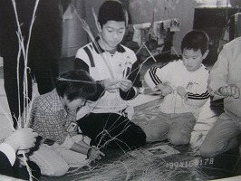 子どもたちが大人に教わりながら藁を使ってしめ縄作りを行っているモノクロ写真