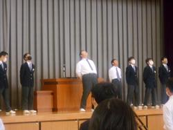 体育館の壇上で、応援団の生徒たちが並び、中央の生徒が体を反らせて声を出している様子の写真