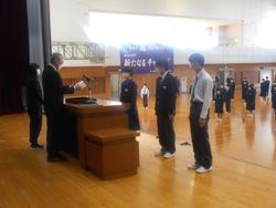 体育館の壇上に上がった3人の生徒が演台に向かって横一列に並んで立ち、演台に立った先生が賞状を読み上げている様子の写真