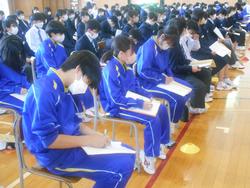 並んだ椅子に座った大勢の生徒たちが、膝に乗せた紙に熱心にメモを取っている様子の写真