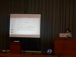 舞台の大きなスクリーンの横にある演台に立ち話をする山内明美先生の写真