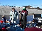 青のコンテナが置かれた側で黒いつなぎを着ている方が魚を両手で持ちその隣で様子をみている男性の写真