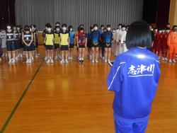 体育館で、紫色のジャージを着た生徒が、部活動ごとに整列したユニフォーム姿の選手たちへ激励の言葉を送っている様子の写真