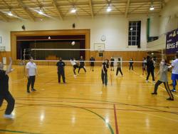 体育館で複数の人々がバレーボールの練習をしており、ボールがネットの上を越えていく瞬間の活気ある様子の写真