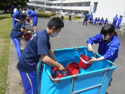 屋外で男子と女子の生徒が水色の水槽から赤いバケツに水を汲んでおり、その奥では並んだ生徒たちがバケツリレーをしている消火訓練の様子の写真