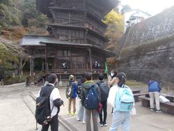 古い木造建築の建物の前で、リュックを背負った児童たちが熱心に説明を聞いている様子の写真