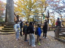 イチョウの紅葉が美しい公園で、緑の旗を持った女性が児童たちに何かを説明している様子の写真