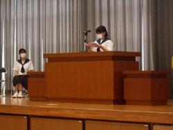 女子生徒が壇上の演台に立ち、原稿を読み上げ、もう一人の女子生徒が横に置かれた椅子に座っている写真