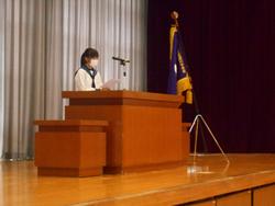 女子生徒が壇上に置かれた旗の横にある演台に立ち、原稿を読み上げている写真