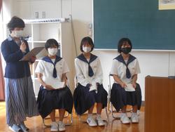 教室で、3人の女子生徒が横並びに椅子に座り、その隣でマイクを持った女性が原稿を読んでいる写真