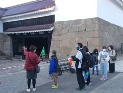 城の石垣と門の前でガイドの説明を聞いている児童たちの写真