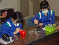 青いジャージを着た女子児童2人が、赤い張り子の牛に絵付けをしている様子の写真