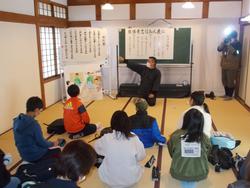 和室で児童たちが畳に座り、黒服の講師が資料を指差し話をしている様子の写真
