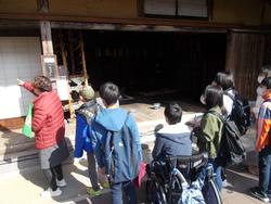 屋外の古民家の前で、ガイドの説明を聞きながら見学している児童たちの様子の写真