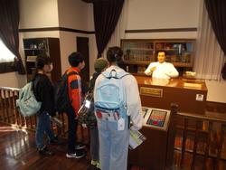 展示スペースで、展示された人形が説明しているように見え、4人の児童がその前で説明を聞いている様子の写真